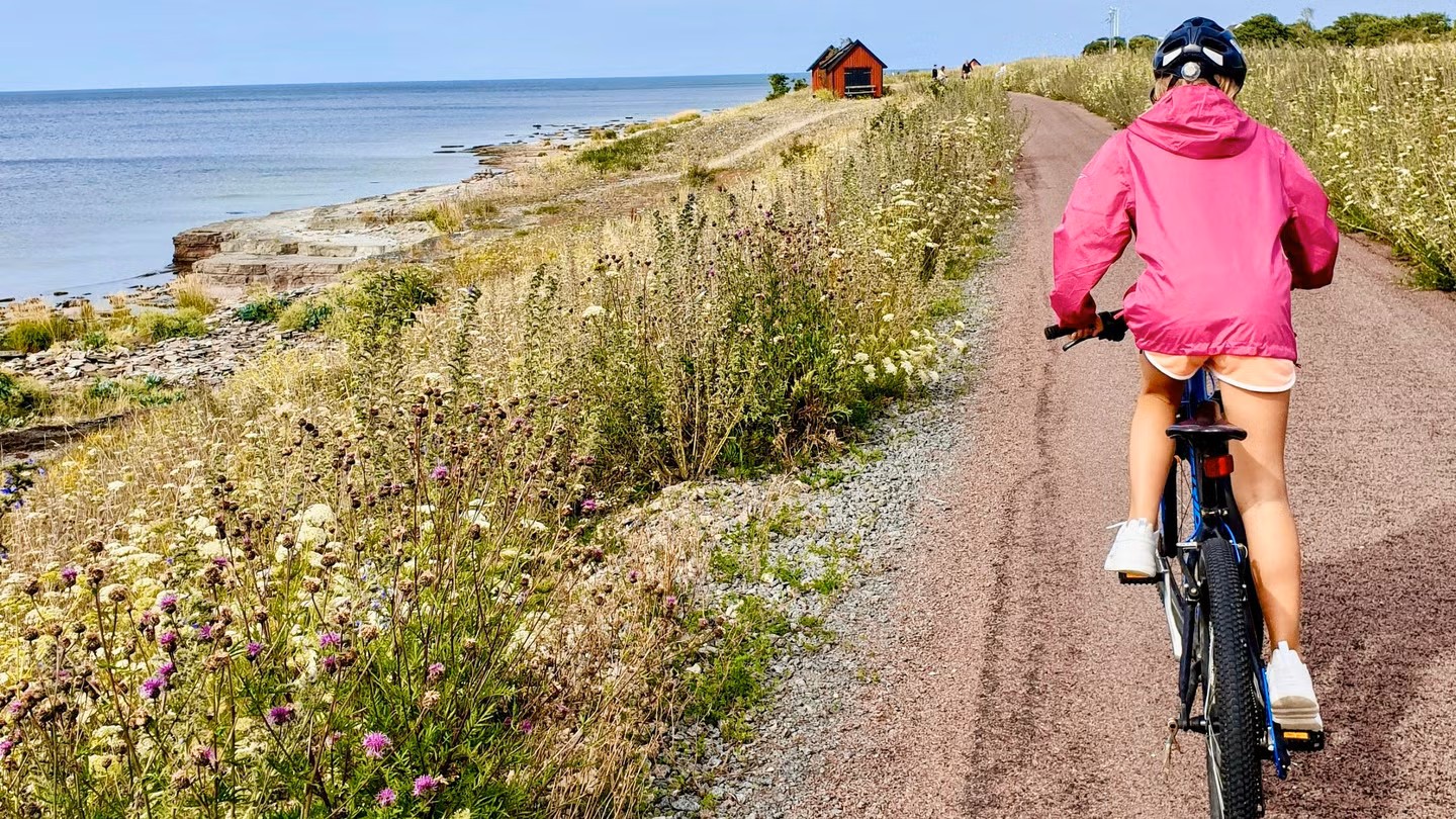 Ett barn med rosa jacka, shorts och cykelhjälm cyklar på en grusväg längs kusten.
