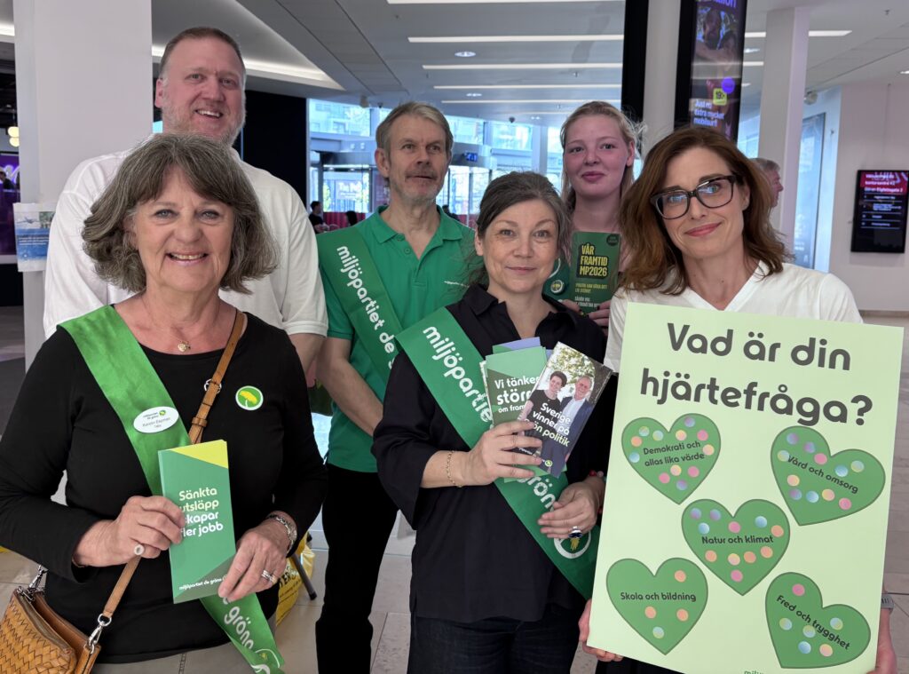 6 medlemmar från Miljöpartiet i Täby står i Täby Centrum och pratar med invånare om partiets politik. Några personer håller upp broschyrer och ett stort plakat med frågan: Vilken är din hjärtefråga?